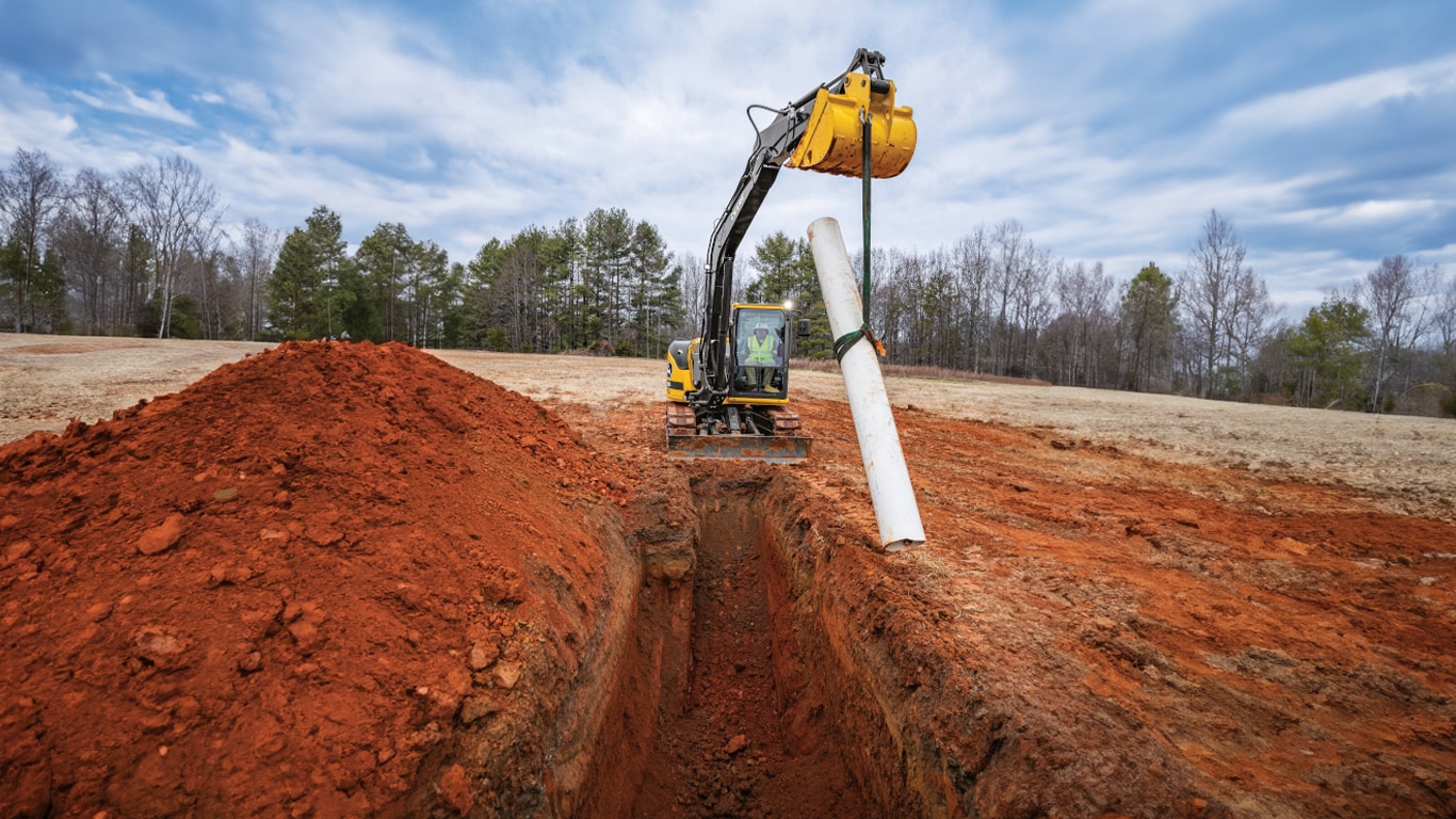 John Deere Excavator laying a pipe into a trench on a worksite.